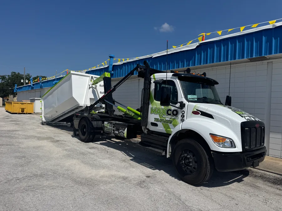 MAXCO hook-lift truck loading a white roll-off dumpster for pickup